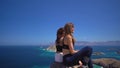 Aerial view of two young tourists sitting on hilltop and admiring landscape of ocean and islands