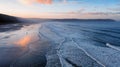 Aerial view of Woolacombe beach and Baggy Point at dawn, waves breaking on a beach where the sunlit clouds are reflected in the