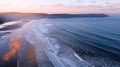 Aerial view of Woolacombe beach at dawn, waves breaking on a beach where the sunlit clouds are reflected in the standing water.