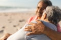 Affectionate biracial senior man sitting with arm around woman at beach during sunny day