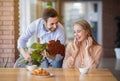 Affectionate millennial man surprising his girlfriend, giving her bouquet of flowers in cafe