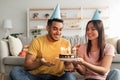 Affectionate millennial multiracial couple in party hats holding birthday cake with lit candles in living room