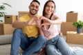 Affectionate multiracial couple making heart gesture with their hands, moving to new house, sitting on floor among boxes