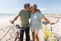 Affectionate multiracial senior couple kissing while standing with cycles at sunny beach against sky