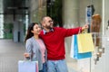 Affectionate young interracial couple standing on street with shopping bags, pointing at store window near city mall Royalty Free Stock Photo