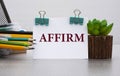 AFFIRM - words on a white sheet with clamps against the background of a cactus and jars with pencils