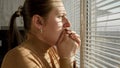 Afraid and scared woman closing her mouth with hand and looking out of the window through blinds Royalty Free Stock Photo