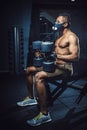 African american athletic man in mask sitting and waiting and looking at himself before lifting dumbbells in front of the mirror. Royalty Free Stock Photo