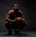 African-American basketball player in sportswear squatting with a ball on a dark background. Royalty Free Stock Photo