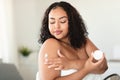 African american bodypositive lady in bath towel applying cream on shoulder, putting on moisturizing body butter at home