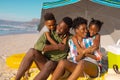 African american boy and girl embracing happy parents sitting on blanket under umbrella at beach Royalty Free Stock Photo