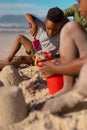 African american boy playing with sand, pail, shovel while sitting with father and sister at beach Royalty Free Stock Photo