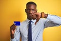 African american businessman holding credit card standing over isolated yellow background with angry face, negative sign showing Royalty Free Stock Photo