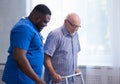African-American caregiver is teaching disabled old man to walk with walker. Professional nurse and handicapped patient