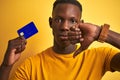 African american customer man holding credit card standing over isolated yellow background with angry face, negative sign showing Royalty Free Stock Photo