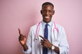 African american doctor man wearing stethoscope standing over isolated pink background smiling and looking at the camera pointing Royalty Free Stock Photo