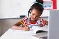African american elementary schoolgirl wearing headphone while writing on book at desk with laptop Royalty Free Stock Photo
