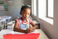 African american elementary schoolgirl writing on book while sitting at desk in classroom Royalty Free Stock Photo