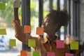 African American employee putting sticker notes to share idea for startup project on glass board in creative office