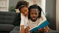 African american father and daughter lying on sofa reading book at home Royalty Free Stock Photo