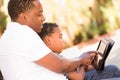 African American Father and Mixed Race Son Using Computer Tablet on Bench in Park Royalty Free Stock Photo