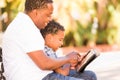 African American Father and Mixed Race Son Using Computer Tablet on Bench in Park Royalty Free Stock Photo