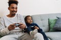 African american father and son playing video game while sitting on couch Royalty Free Stock Photo