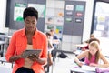 African american female teacher using tablet in classroom with diverse pupils, with copy space