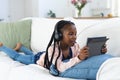 African american girl lying on couch, listening to music and using tablet at home, copy space Royalty Free Stock Photo