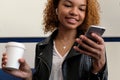 African American girl smiling, looking into her phone. A white glass with coffee in hand. A beautiful young modern black woman, in