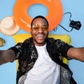 African American guy in funky outfit lying on lilo, taking selfie during beach vacation on blue background, top view Royalty Free Stock Photo