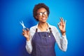 African american hairdresser woman holding scissors over blue background doing ok sign with fingers, excellent symbol