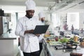 African american head chef with laptop searching for dish recipes on internet while standing in restaurant kitchen. Royalty Free Stock Photo