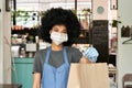 African American female cafe worker wearing face mask holding takeaway food bag.