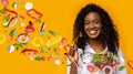 African American lady with bowl of fresh summer salad and flying vegetables on orange background, collage Royalty Free Stock Photo