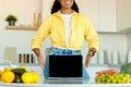 African american lady showing laptop with blank screen at table with organic vegetables at kitchen interior, mockup