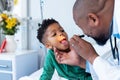 African american male doctor examining mouth of boy patient with penlight in hospital Royalty Free Stock Photo