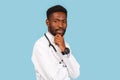 African american male doctor with stethoscope holds hand to chin and thinks over blue background Royalty Free Stock Photo