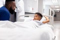 African american male doctor talking to boy patient in hospital bed in corridor with copy space Royalty Free Stock Photo