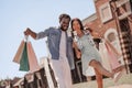 African american man and asian woman with shopping bags and taking selfie with smartphone using selfiestick outdoors at daytime