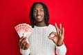 African american man with braids holding 20 israel shekels banknotes doing ok sign with fingers, smiling friendly gesturing