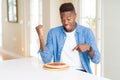 African american man eating pepperoni pizza at home very happy pointing with hand and finger Royalty Free Stock Photo