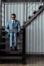 African american man in glasses using laptop while sitting on stairs in office