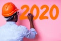 African American man with helmet hardhat white on pink wall with brush tool drawing new year 2020 paint orange color