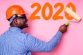 African American man with helmet hardhat white on pink wall with roller brush tool drawing new year 2020 paint orange