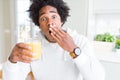 African American man holding and drinking glass of orange juice cover mouth with hand shocked with shame for mistake, expression Royalty Free Stock Photo