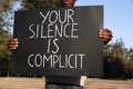 African American man holding sign with phrase Your Silence Is Complicit outdoors, closeup. Racism concept