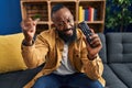 African american man holding television remote control annoyed and frustrated shouting with anger, yelling crazy with anger and Royalty Free Stock Photo