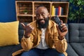 African american man holding television remote control pointing thumb up to the side smiling happy with open mouth Royalty Free Stock Photo