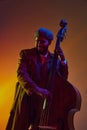 African-American man, musician in vintage coat and cap creates rhythm on bass in neon light against gradient background.